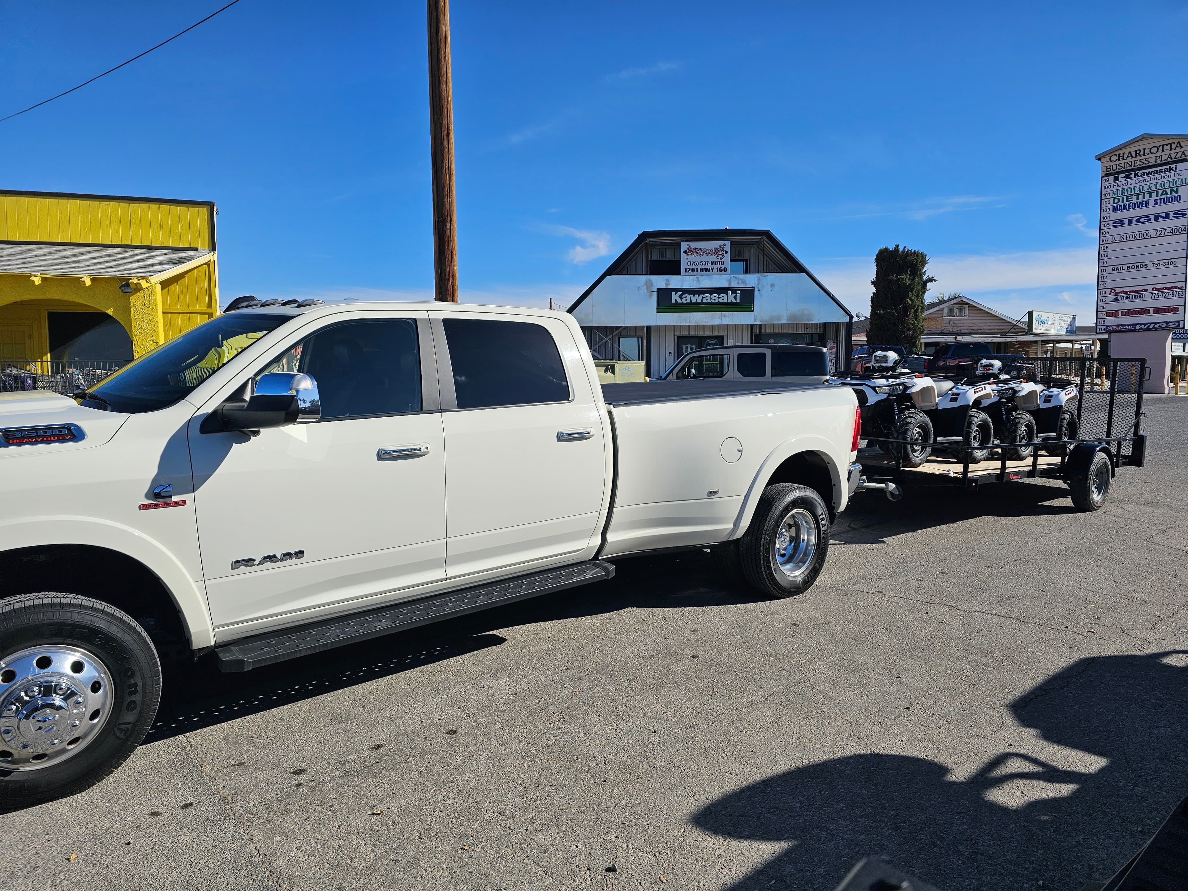 White Ram 3500 dually pickup towing a utility trailer loaded with ATVs outside a Kawasaki dealership